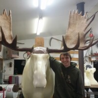 A person stands next to a large mounted moose head in a workshop filled with tools and animal sculptures.