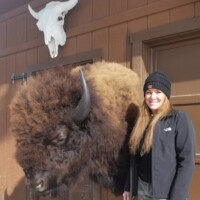 A woman stands next to a mounted bison head on a wooden wall with a cow skull displayed above.