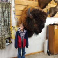 A smiling child stands indoors next to a mounted bison head displayed on a wooden wall.