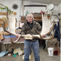 Older man in a workshop holding a large pair of moose antlers, with tools and shelves visible in the background.